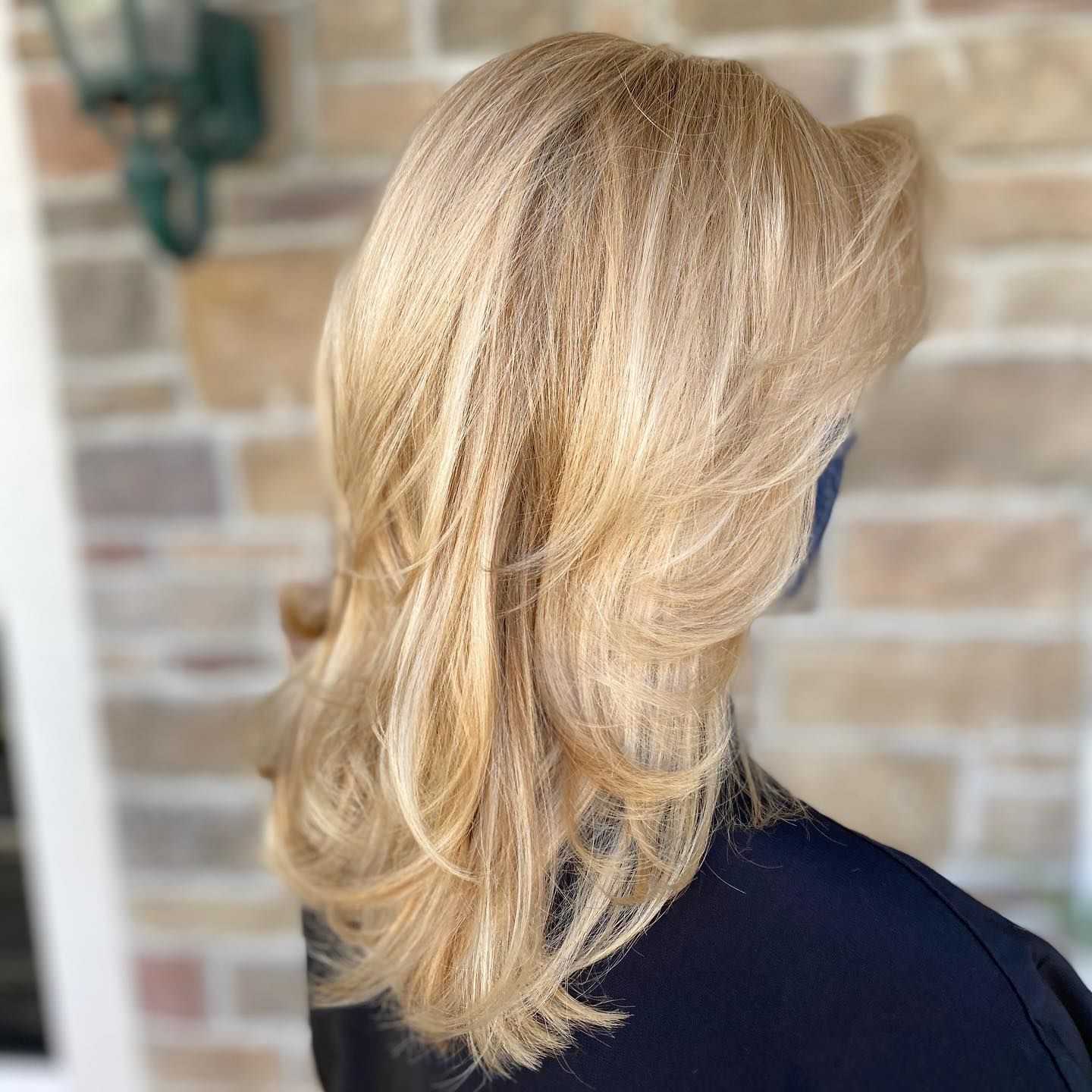 Blonde hair with soft waves and layers against a brick wall background.